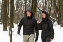 A man and a woman walking through the snowy woods, each wearing a black hooded Gamma graphene jacket.