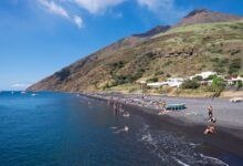 The island of Stromboli, with dozens of people sunning themselves on a black sand beach and playing in the blue water, the volcano rising behind them.