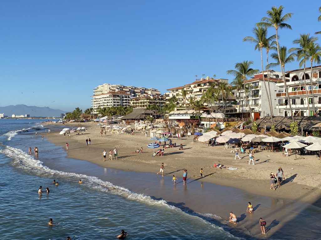 A beach lined with tall white apartment buildings and palm trees, lots of people playing in the water.