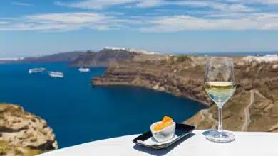 En el primer plano de esta imagen hay una copa de vino blanco con la caldera de Santorini al fondo. Santorini produce algunos de los mejores vinos griegos.