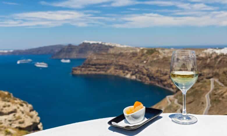 En el primer plano de esta imagen hay una copa de vino blanco con la caldera de Santorini al fondo. Santorini produce algunos de los mejores vinos griegos.