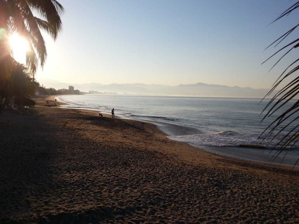 A man and his dog walking along an empty beach at sunrise. Mountains in the background.