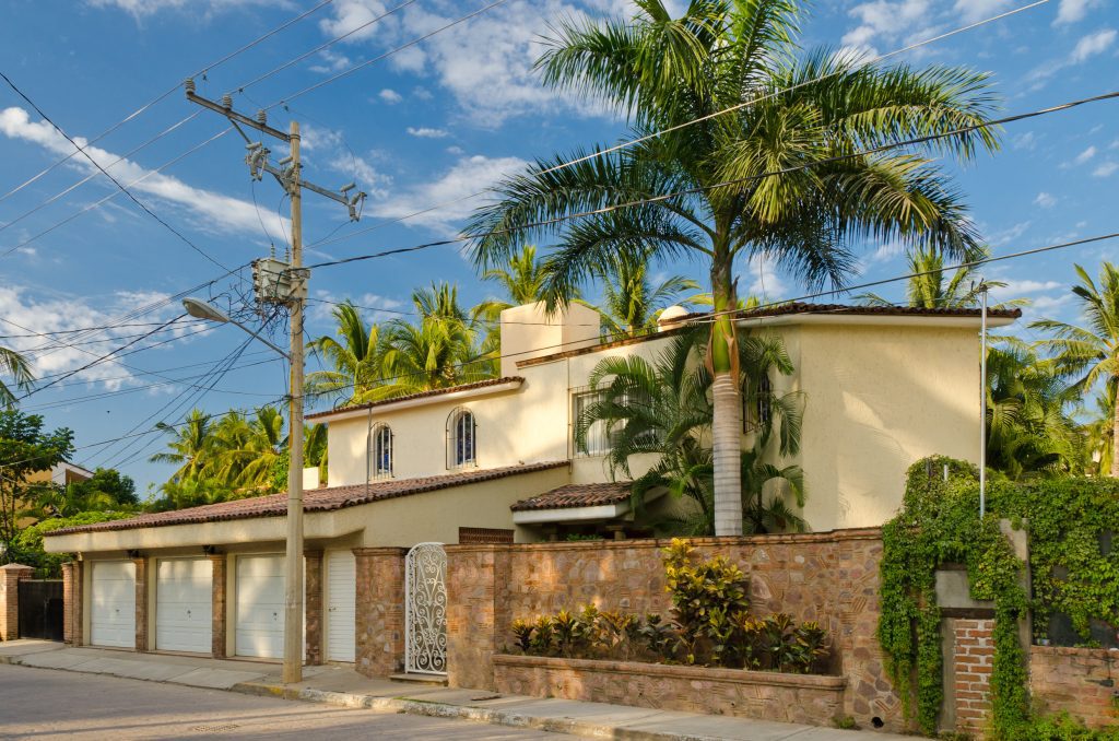 A luxury home with a three-car garage behind a stone fence, surrounded by palm trees.