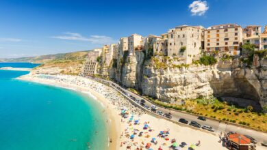 Tan stone buildings built into the cliffs of Tropea, overlooking a long white beach and bright clear turquoise water.