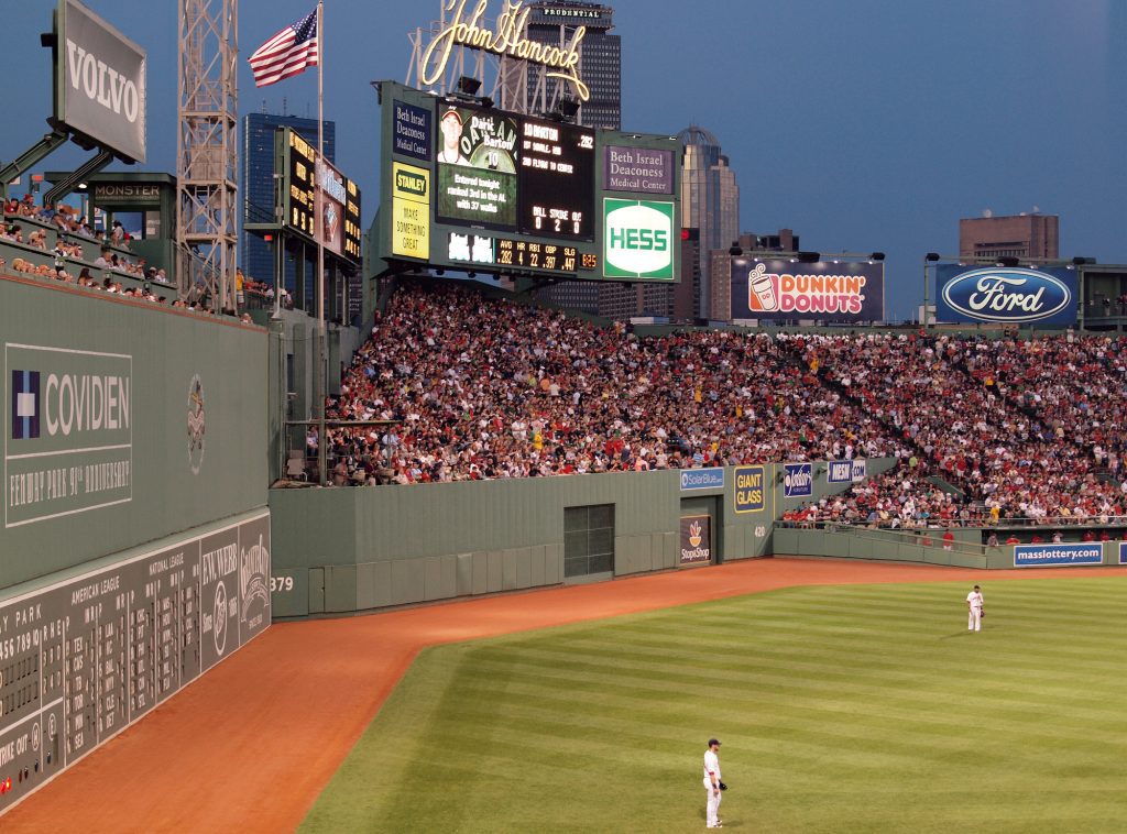 A view of the outfield at Fenway Park baseball stadium, people filling the stands and some people sitting in the seats atop the Green Monster (tall green wall).