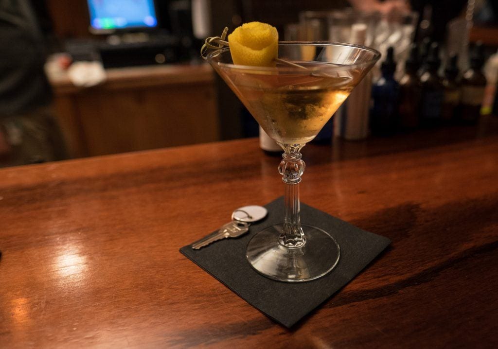 A cocktail in a martini glass on a wooden bar, a lemon rind hanging off the edge.