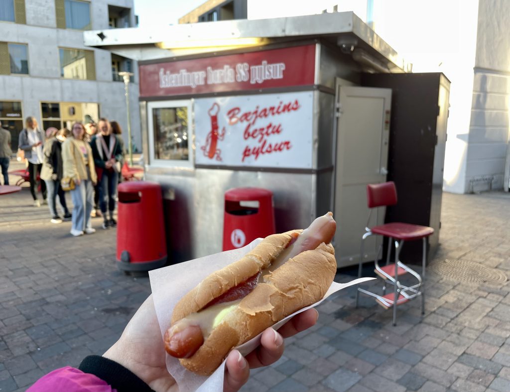 A hand holds up a hot dog covered in various sauces in front of a metal and red and white little kiosk selling hot dogs. Lots of people waiting in line.