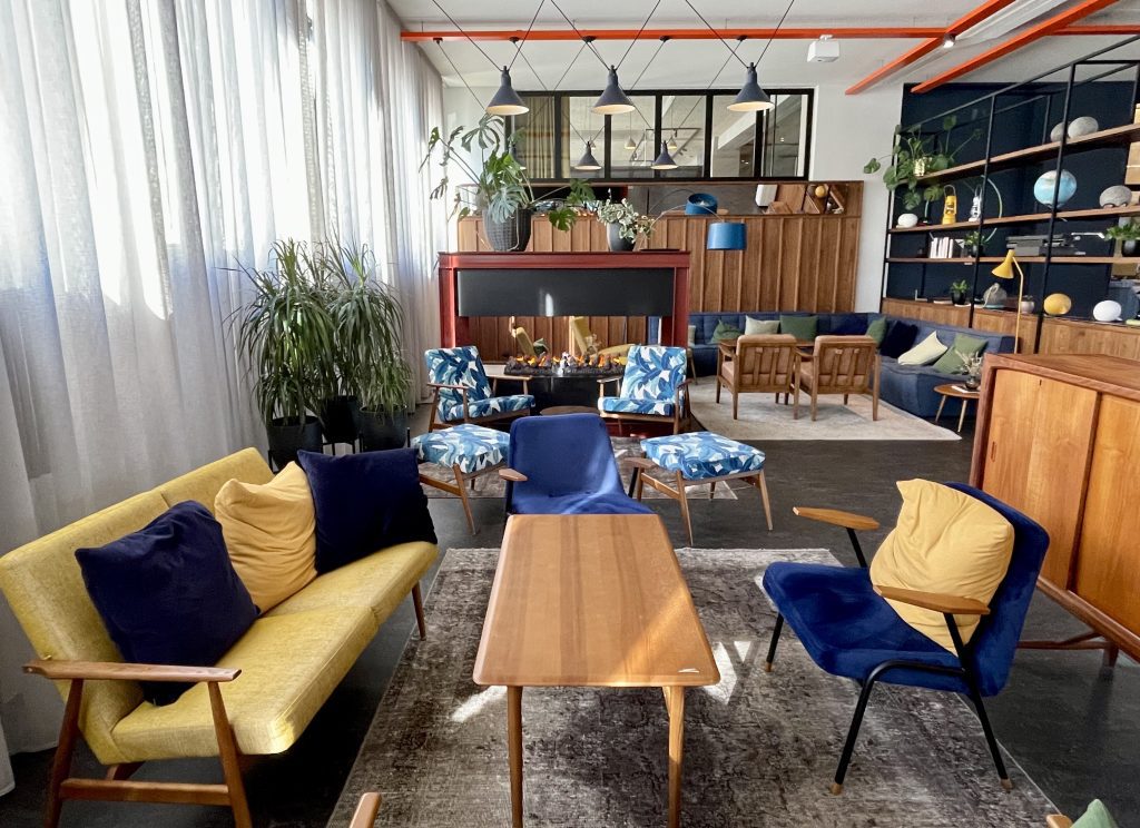 A cozy hotel lobby with mid-century modern sofas and chairs in shades of yellow and navy, leading to an open fireplace surrounded by more chairs.
