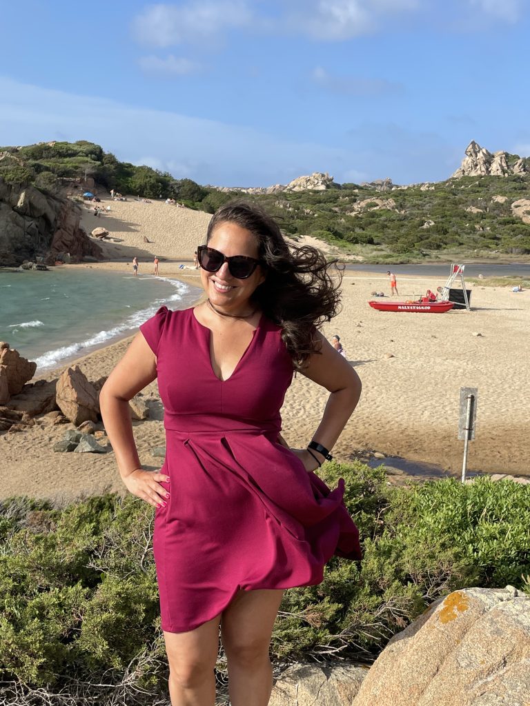 Kate smiles, hands on hips in her burgundy short dress, in front of a beach in Sardinia. It's windy and her hair and dress are flying.