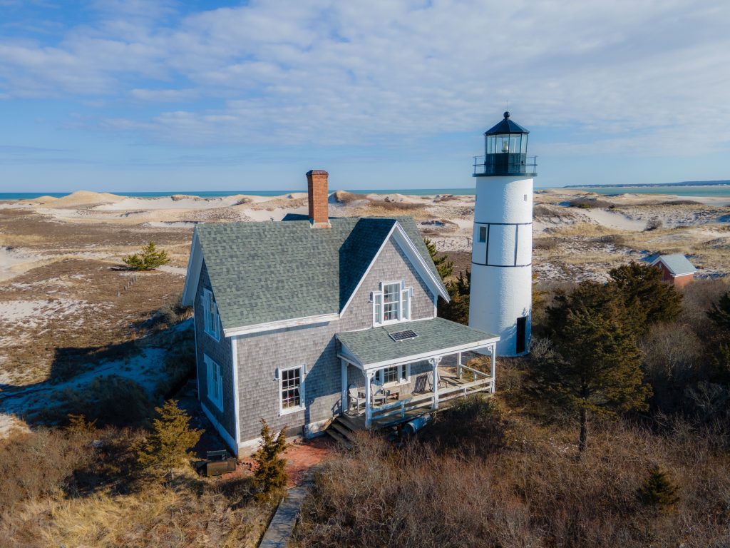A gray clapboard house in a beach next to a white lighthouse.