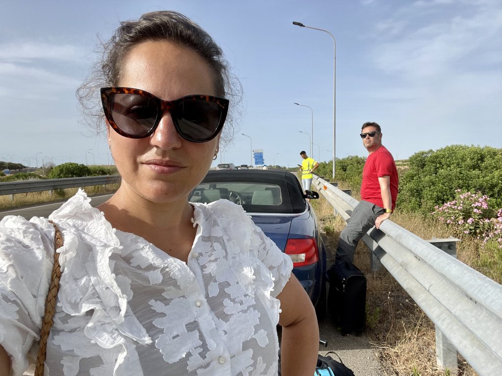 Kate takes a glaring selfie at the camera wearing a frilly white button-down blouse and sunglasses. She's standing on the highway. Behind her is a blue car and Charlie sitting on the guardrail, angrily glaring at the camera. Behind them is a mechanic in a yellow shirt.