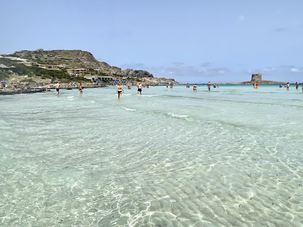 Beautiful clear pale green water at a beach in Sardinia. Several people are in the water and you can see it doesn't even get knee deep in the distance.