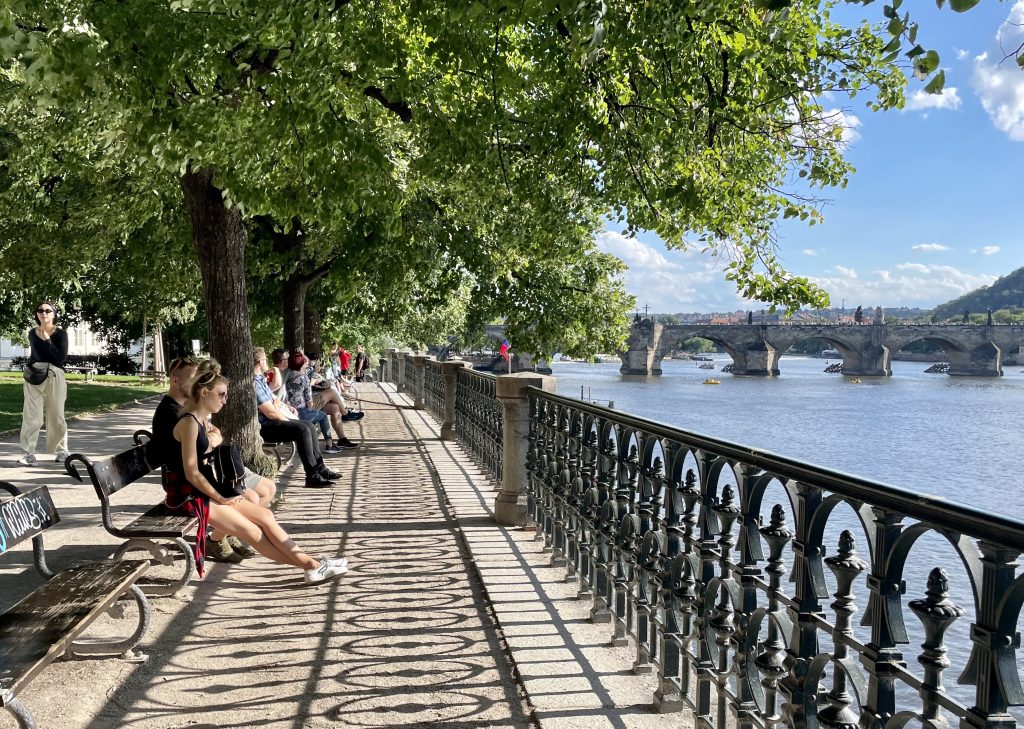 People sitting in front of wrought-iron benches in front of the river in Prague, the iron casting shadows in front of them.