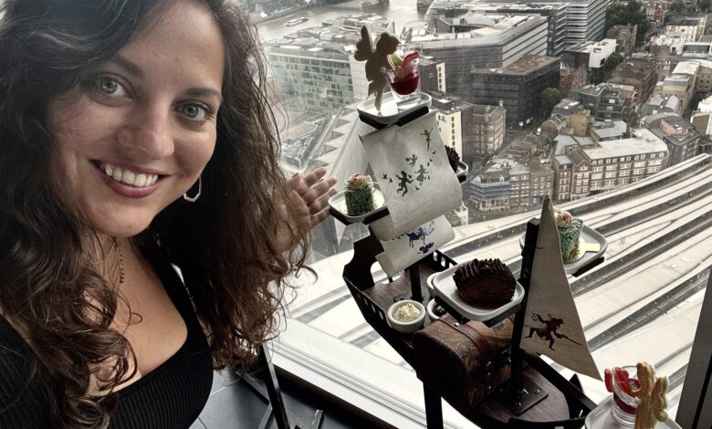 Kate smiling in front of a small pirate ship topped with afternoon tea pastries. Behind her is the city skyline of London, including the Tower Bridge.