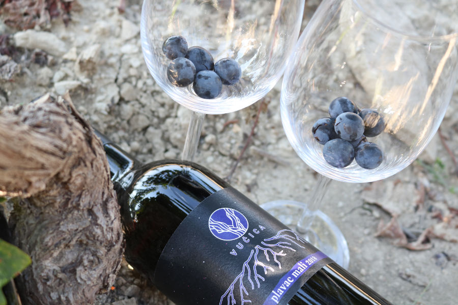 Esta imagen muestra una botella de vino Vucica y dos copas de vino vacías con uvas negras. 