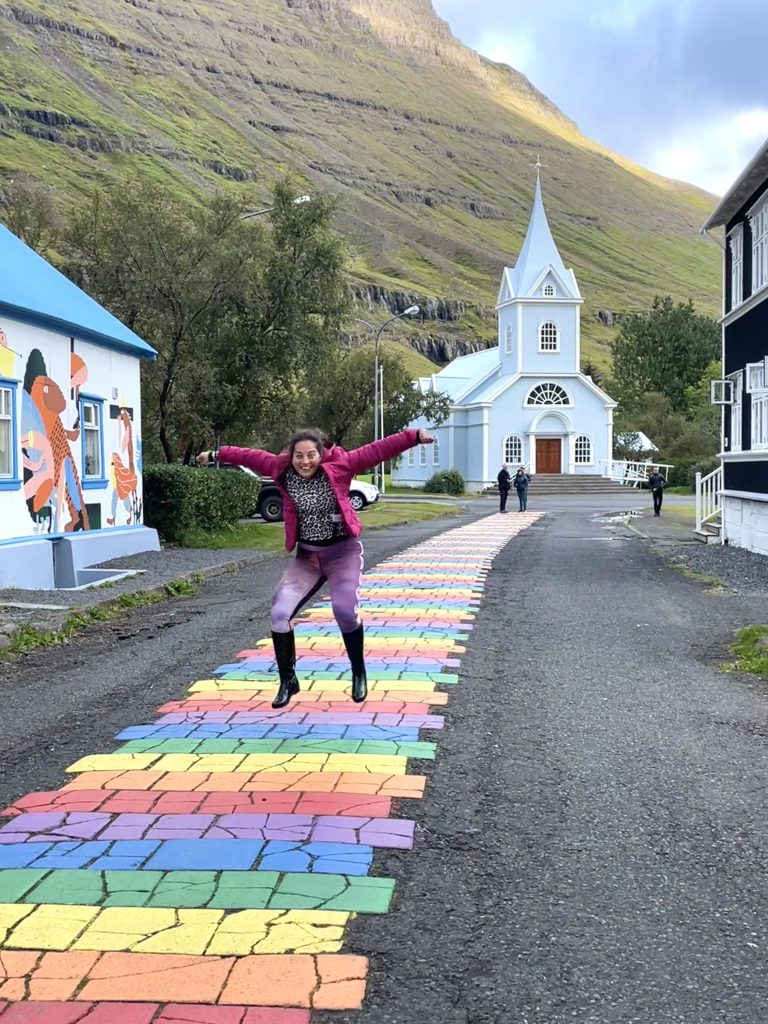 Kate jumping into the air like a goofball, wearing a pink coat, purple leggings, and tall black boots. She's on a rainbow-striped street and behind her is a pale blue church, followed by a steep green mountain.
