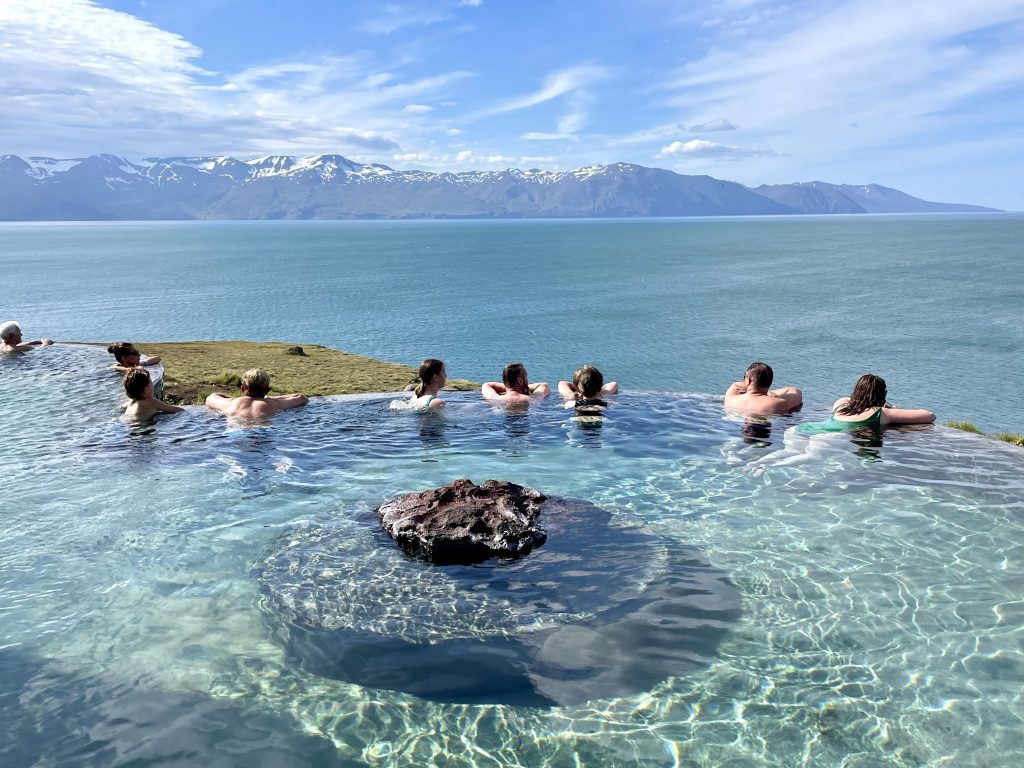 Several people sitting in a hot thermal spring overlooking the ocean and snow-covered mountains in the distance.