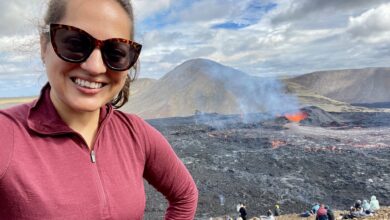 Kate takes a smiling selfie in sunglasses and a red pullover. Behind her is a smoking volcano with a crater full of bubbling red lava. You can see a bunch of people sitting on a hill watching the volcano spew.