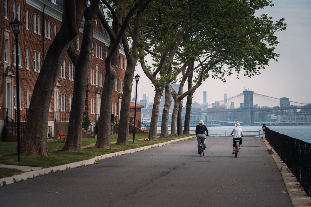 Two bikers riding down a path, to the left is a building and beautiful trees and in front is water and the NYC skyline