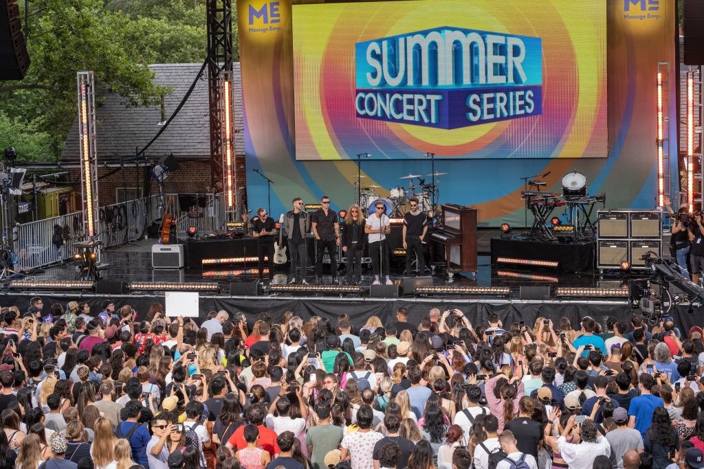 The band One Republic standing on a stage in front of a crowd, the words Summer Concert Series behind them.