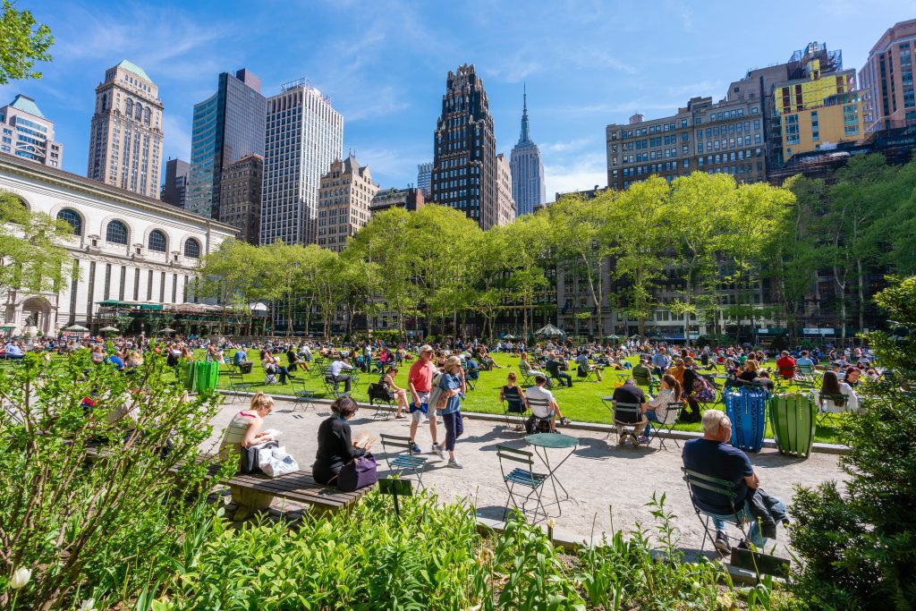 People gathered on benches and the green space in a park, with the New York skyline behind them