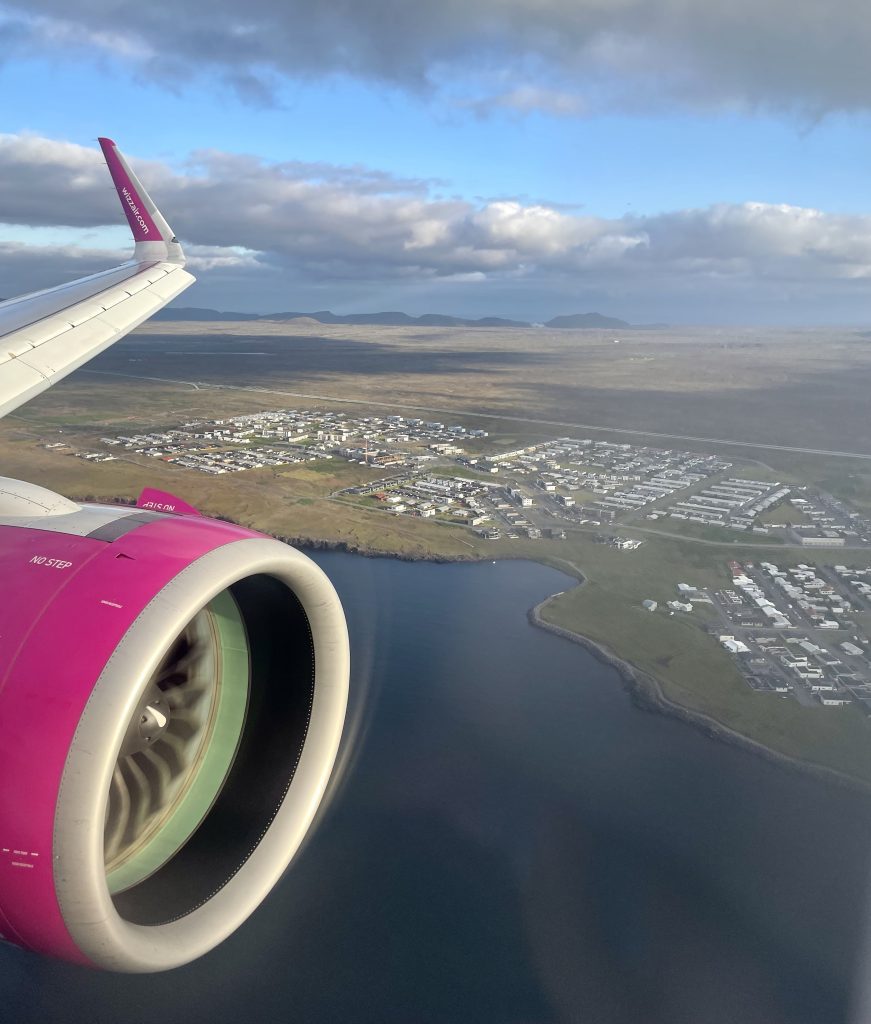 The view out the plane window, you can see a wing and engine flying over a small flat town on the Icelandic coast.