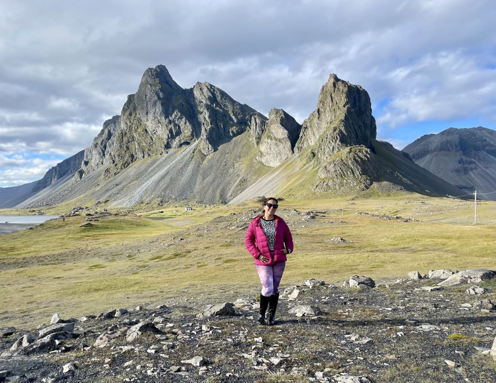 Kate smiles in a pink jacket, purple leggings and black boots, standing in front of a jagged gray-green mountain on a sunny day.