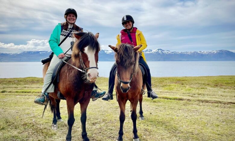 Amanda and Kate on horseback, side by side. The horses are short and brown with shaggy hair, and behind them is a calm bay and snow-covered mountains.