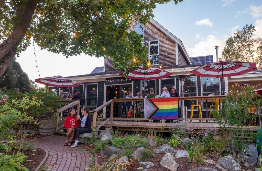 A big restaurant in a shingled house with a big porch, a Pride flag, and people sitting outside and enjoying beers.