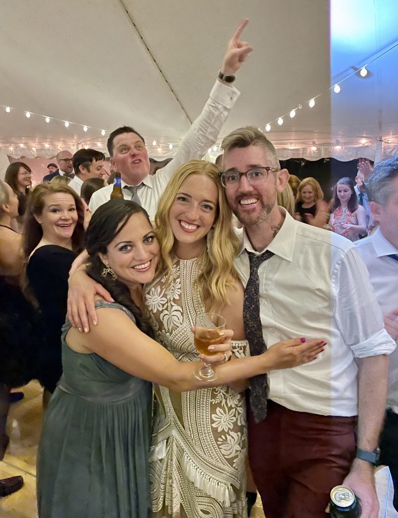 Kate in a long gray dress with her arms around her sister, with blonde wavy hair and a white embroidered dress, and Matt in a white shirt and dark tie. They are on a dance floor at a wedding.
