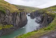 A canyon lined with gray columns of rock, with a turquoise river flowing through it. The sides are grassy and there