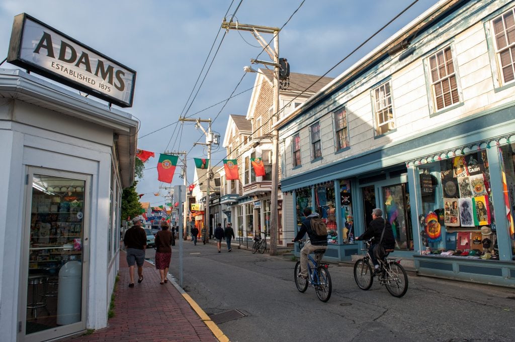 People riding bikes down a pedestrianized street in Provincetown surrounded by white clapboard houses.