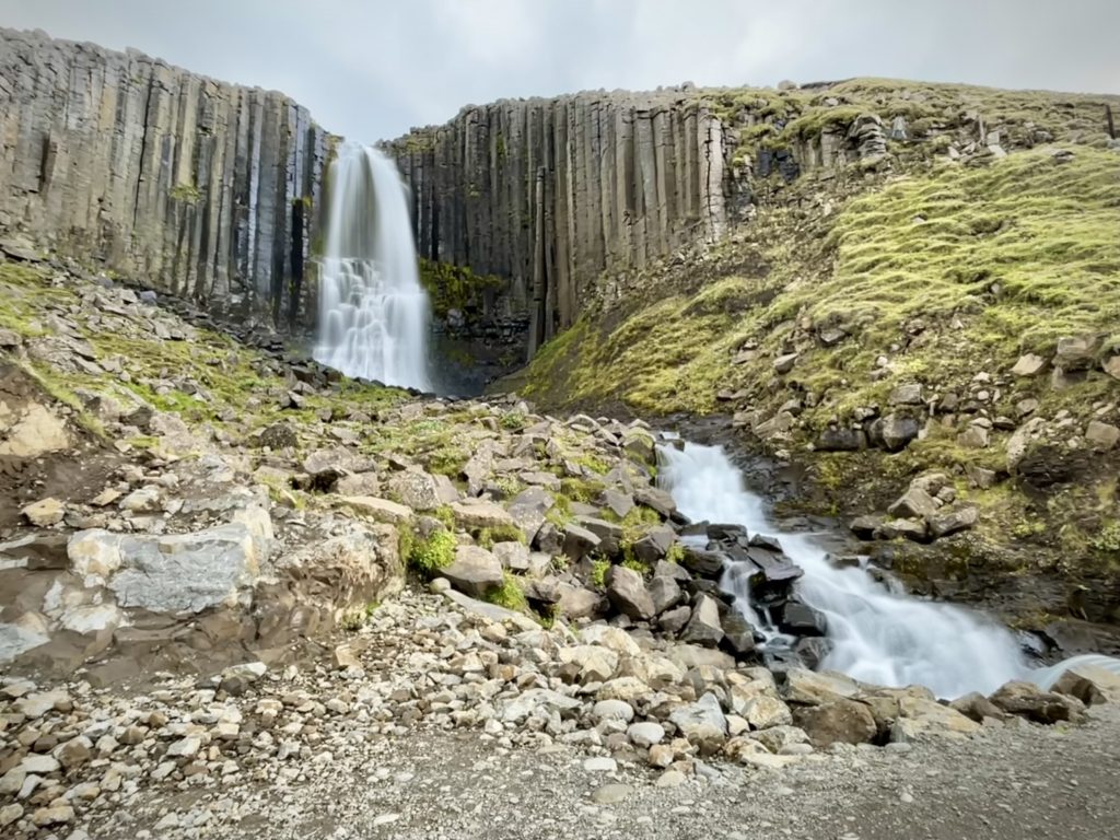 A waterfall emerging from a wall of long, skinny, vertical stone columns.