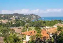 A view of hills topped with palm trees and orange-roofed villas leading to the bright blue ocean.