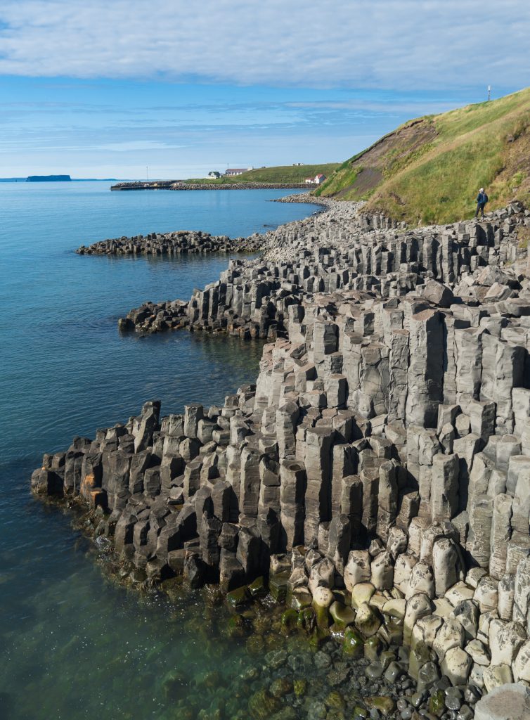 A coastline of tall gray basalt columns next to the calm blue sea.
