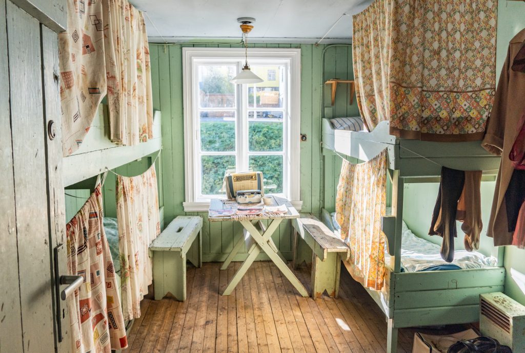 An old-fashioned girls' dorm with four bunk beds, patterned sheets hanging in front of each one, and a 50s radio on the table.