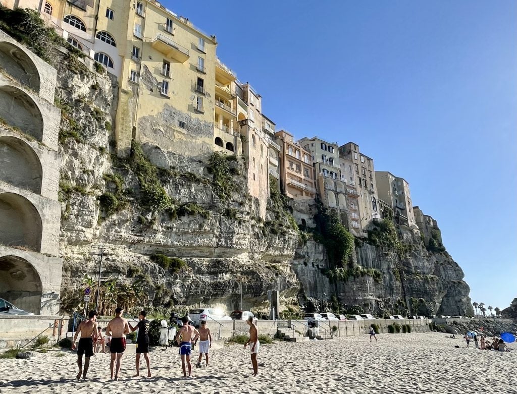 A group of teenage boys gathered on the beach in Tropea, a tall cliff behind them.