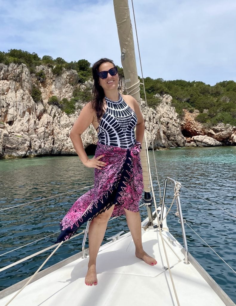 Kate standing on the front of a sailboat, wearing a black and white bathing suit and a pink sarong as a skirt. Behind her is bright blue water and some rocky cliffs.