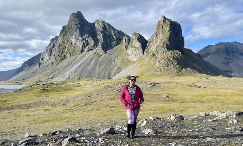 Kate standing in front of a craggy green mountain in Iceland. She wears a pink jacket and has her hands in her pockets, grinning.