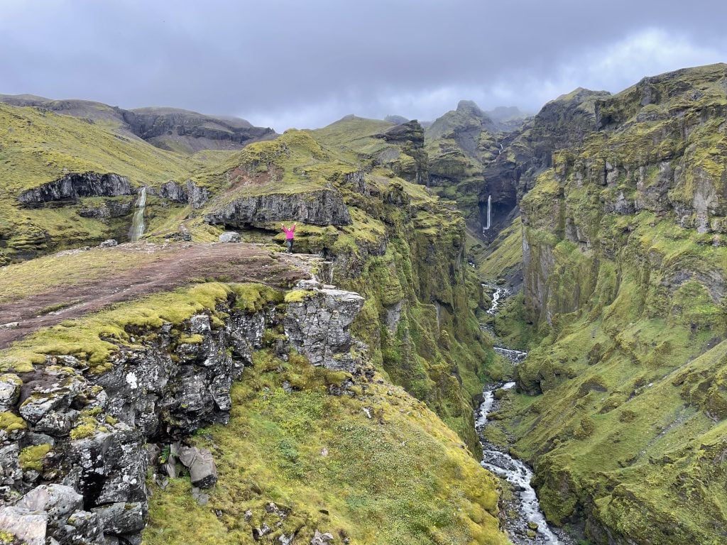 A misty green canyon filled with skinny waterfalls and a narrow river. Kate stands in the distance in a hot pink jacket, holding her arms up in triumph.