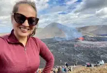 Kate taking a smiling selfie in sunglasses in front of an active volcano spewing bright red lava.