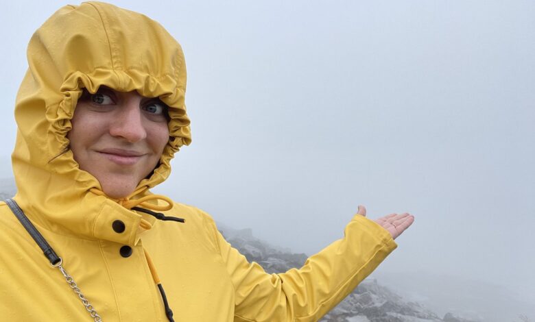 Kate standing in a hooded yellow raincoat, holding her hand out toward a foggy landscape where you can see almost nothing behind her.