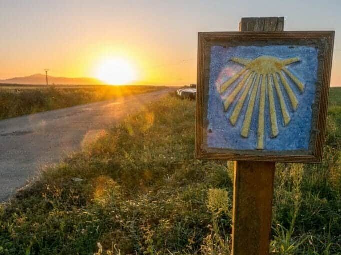 Waymarker of Camino de Santiago Yellow shell on blue background next to a road during sunset - Learn more about the 7 most popular Camino de Santiago Routes to Santiago de Compostela - the most popular pilgrimage in the world.