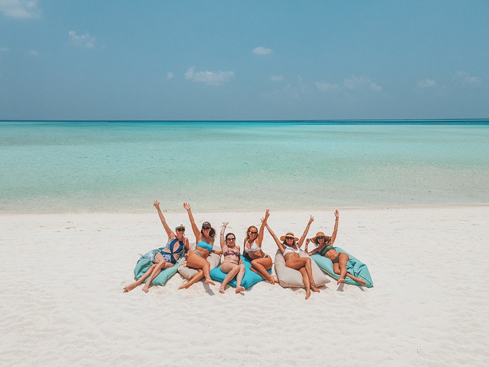 Señoras con los brazos levantados, sentadas en tumbonas en la playa junto al agua azul clara, Maldivas