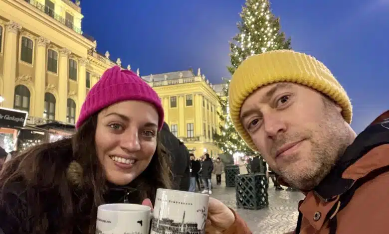 Kate and Charlie toasting gluhwein mugs with hats on in front of a bright yellow palace with a Christmas tree.
