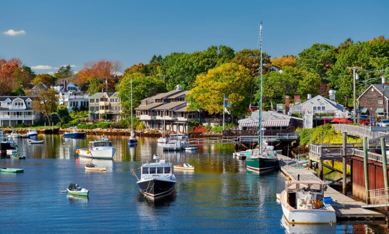 Fishing boats docked in a smooth harbor in front of waterfront homes in Ogunquit, Maine. In the background are trees just starting to turn red and yellow.