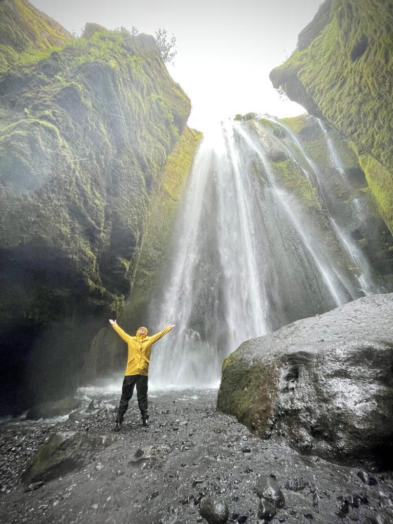 Kate standing in a cave with a waterfall falling through it, wearing a yellow raincoat and holding her arms up in joy.