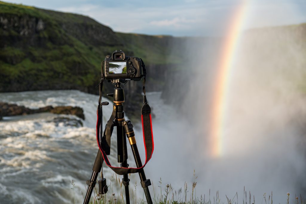 A camera on a tripod in front of an Iceland waterfall with a rainbow.