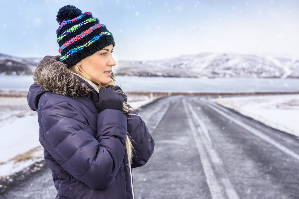 A woman in a winter coat and hat stands on a snowy Icelandic road.