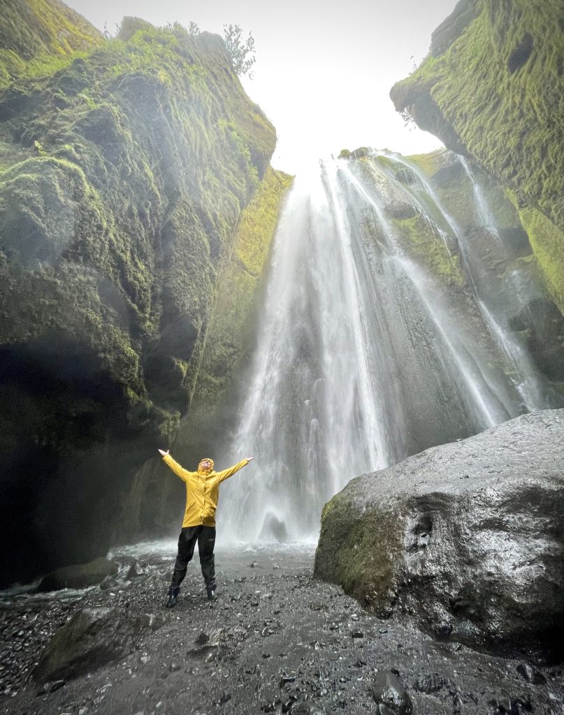 Kate standing next to a thundering waterfall wearing a yellow hooded raincoat.